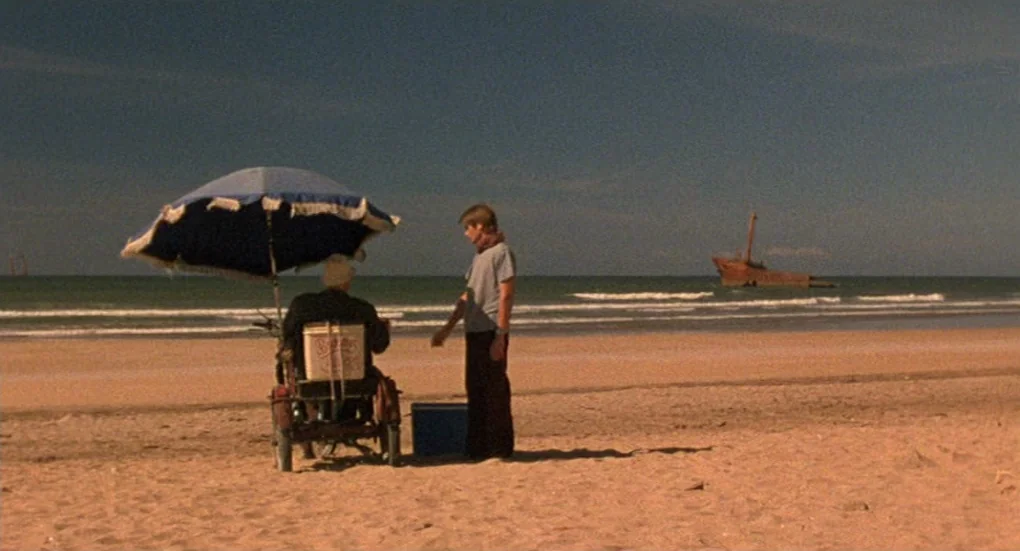 Furia (1999) movie still: wide — A vast sandy beach stretches toward the ocean with a rusted shipwreck in distance. Two men interact near a blue…