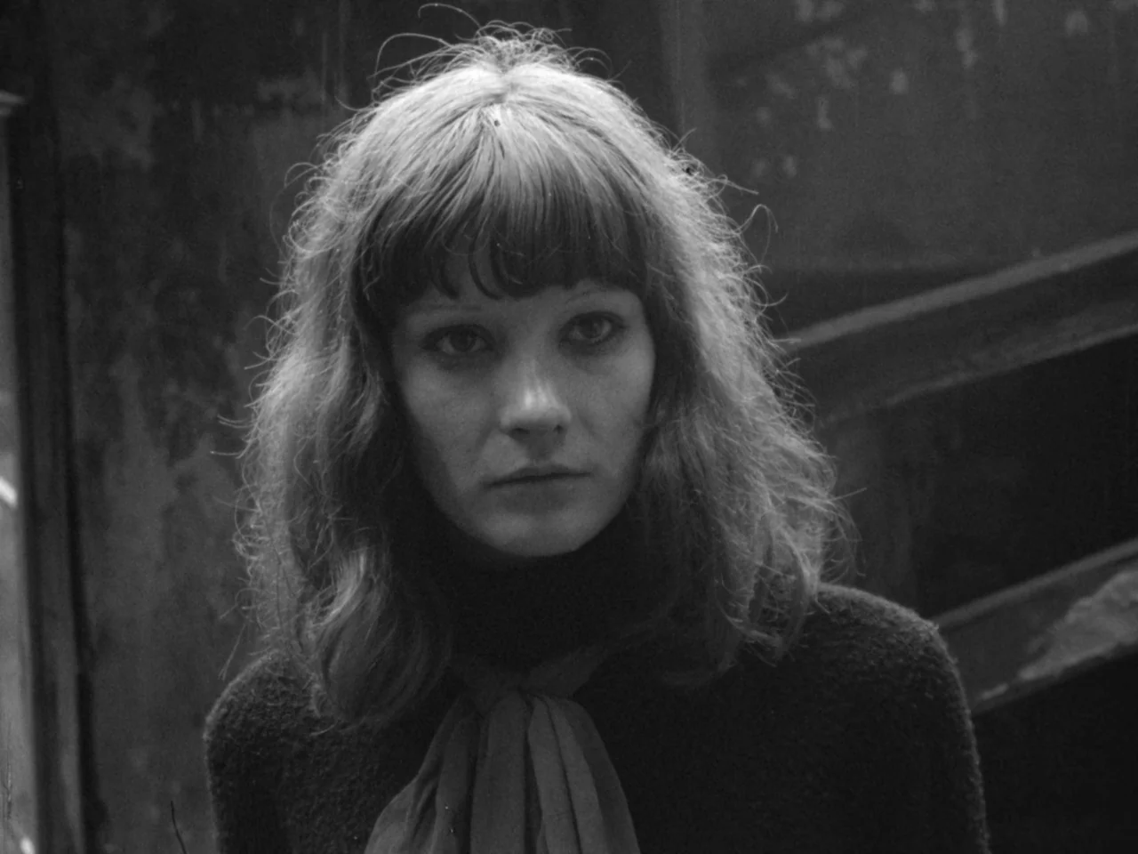 A Woman Kills (1968) movie still: closeup — A woman stands in a dimly lit, weathered alleyway with a textured wall behind her. She faces forward,…