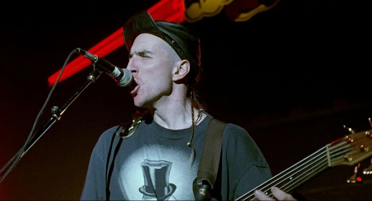Bill and Ted’s Bogus Journey (1991) movie still: medium — A dark concert venue with a red glowing neon sign in the background. A male bassist with…