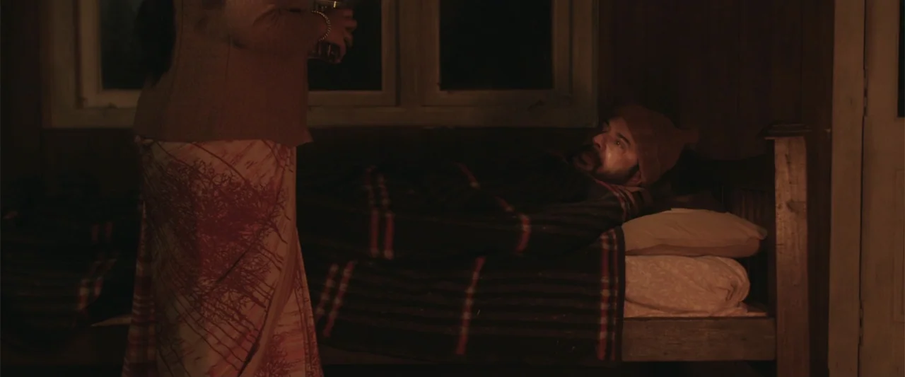 Peranbu (2019) movie still: medium — A dimly lit room contains a wooden bed with a plaid blanket and patterned pillows. A man rests under the…