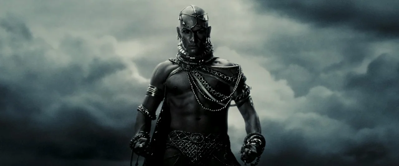 300 (2006) movie still: medium — A muscular man stands against a dark, stormy, overcast sky. He is adorned in extensive metal jewelry and chest…