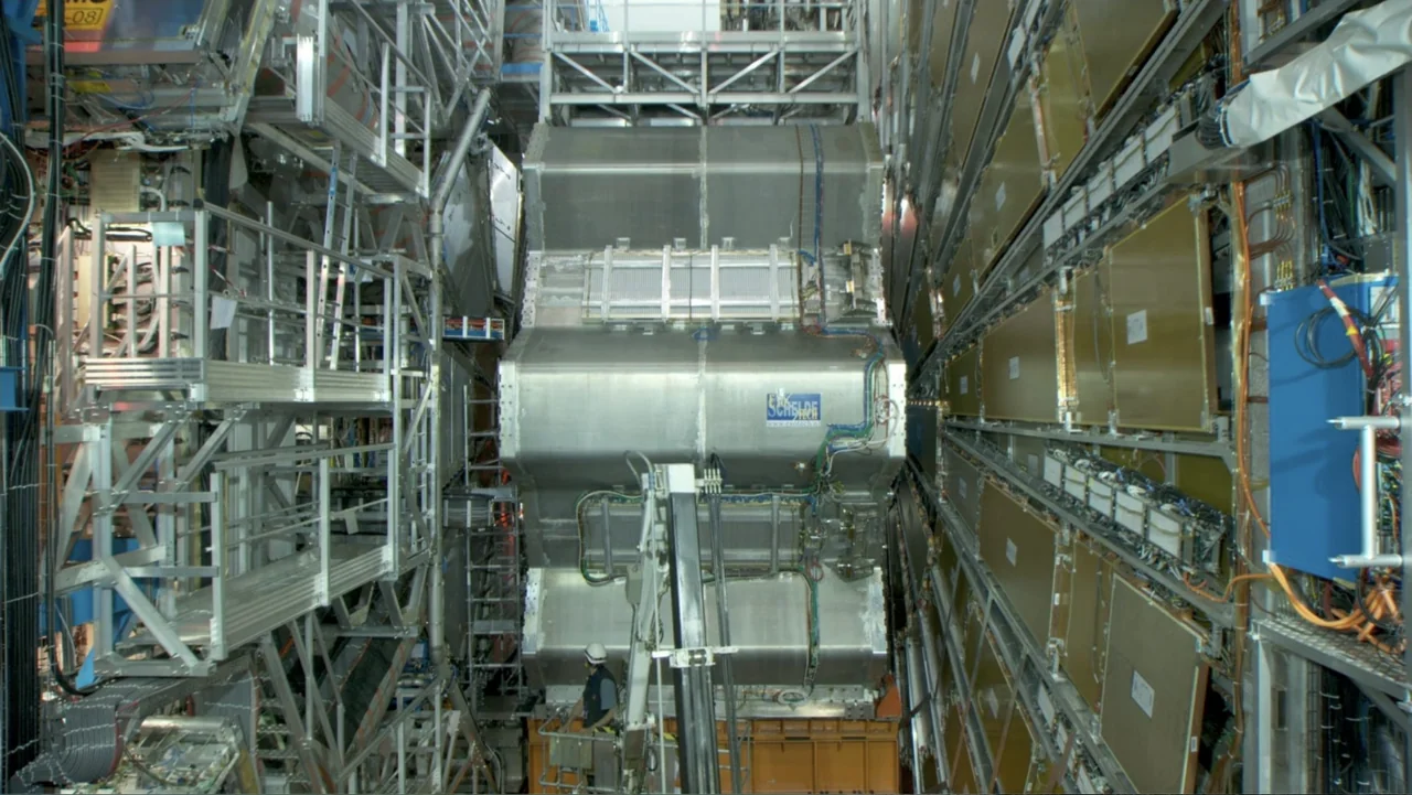 CERN (2013) movie still: wide — A massive particle accelerator detector sits inside a large laboratory facility with complex scaffolding. A single…