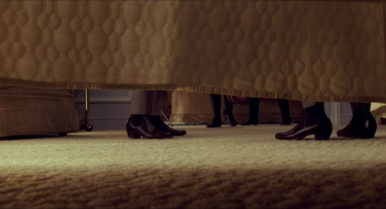 I Wanna Hold Your Hand (1978) movie still: wide — A low-angle view shows the legs and feet of people standing in a room. A textured beige bedspread…