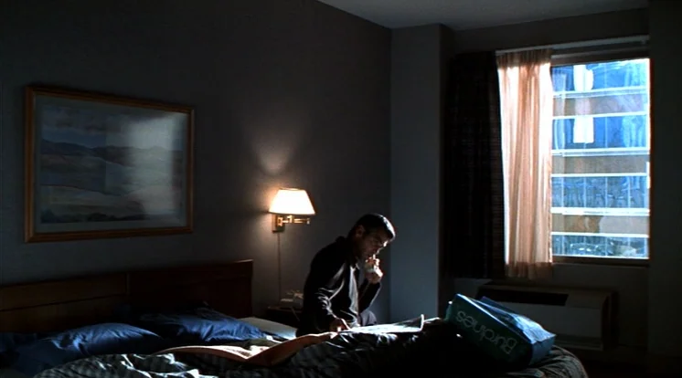 Out Of Sight (1998) movie still: wide — A dimly lit hotel room contains a bed, a wall lamp, and a window. A man sits on the edge of the bed looking…