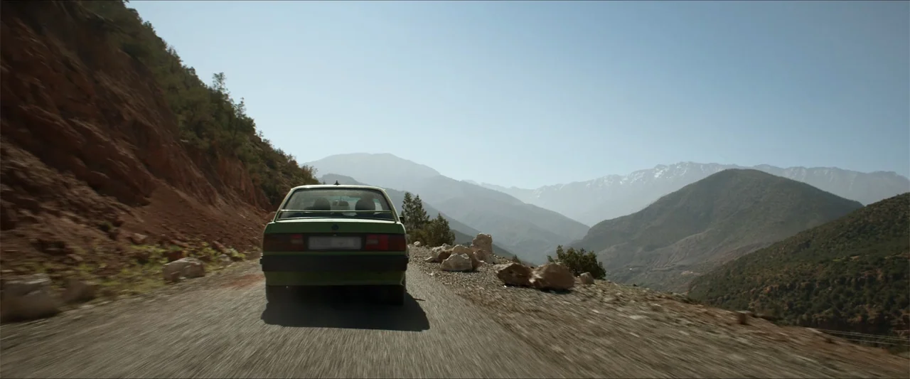 An Endless Wedding (2025) movie still: wide — A green sedan drives along a narrow, unpaved road cut into a steep mountain slope. The car moves…