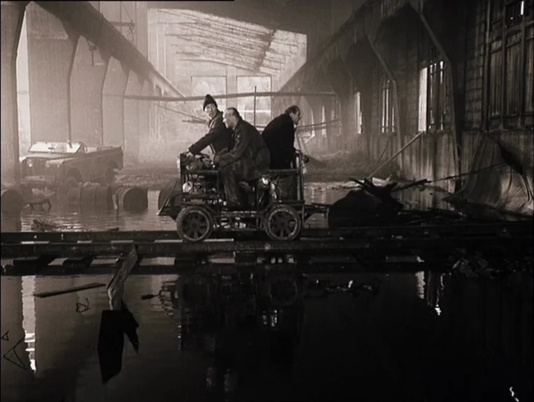 Stalker (1979) movie still: wide — A desolate, flooded industrial site features rusted metal structures and stagnant water. Three men ride a small…