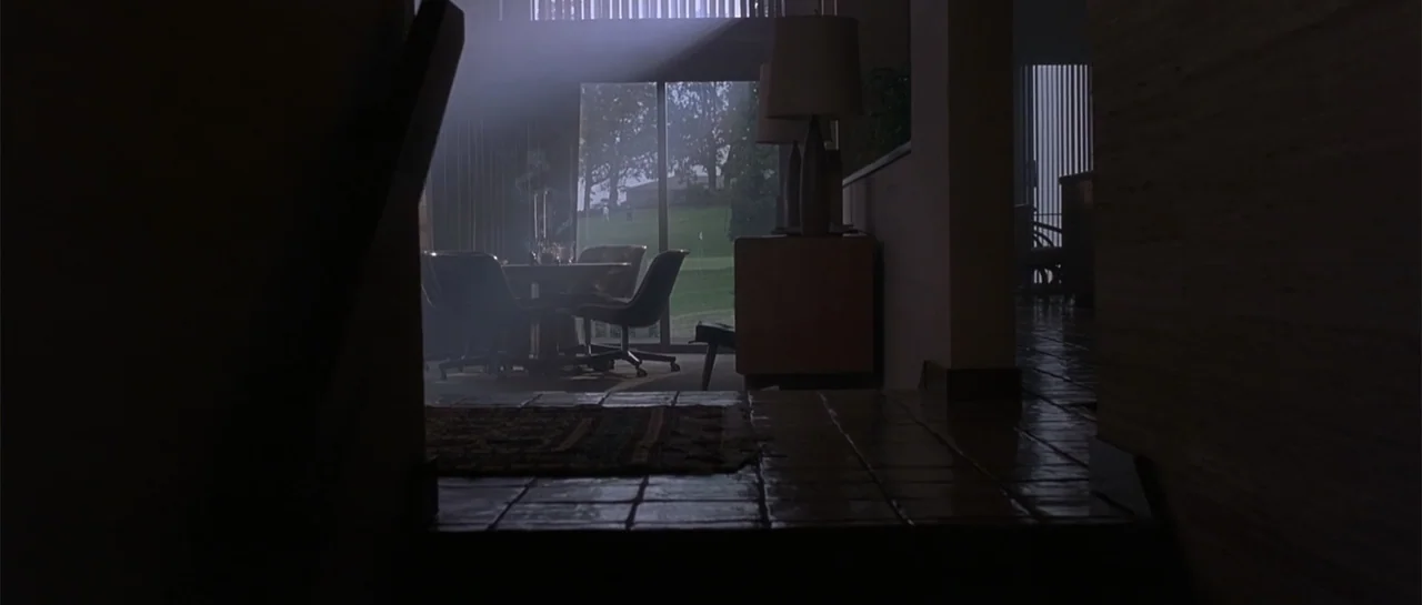 Bandits (2001) movie still: wide — A dimly lit residential interior features dark tile flooring and mid-century modern furniture pieces. A glass…