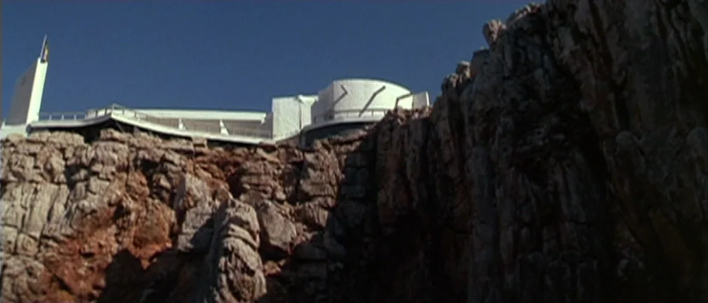 Boom! (1968) movie still: establishing — A white modern structure sits atop a steep, rugged rock formation overlooking the sea. The architecture has…