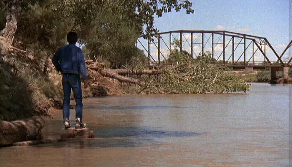 Badlands (1973) movie still: fullbody — A man in denim stands on a rock by a muddy riverbank. A metal truss bridge spans the river in the background…