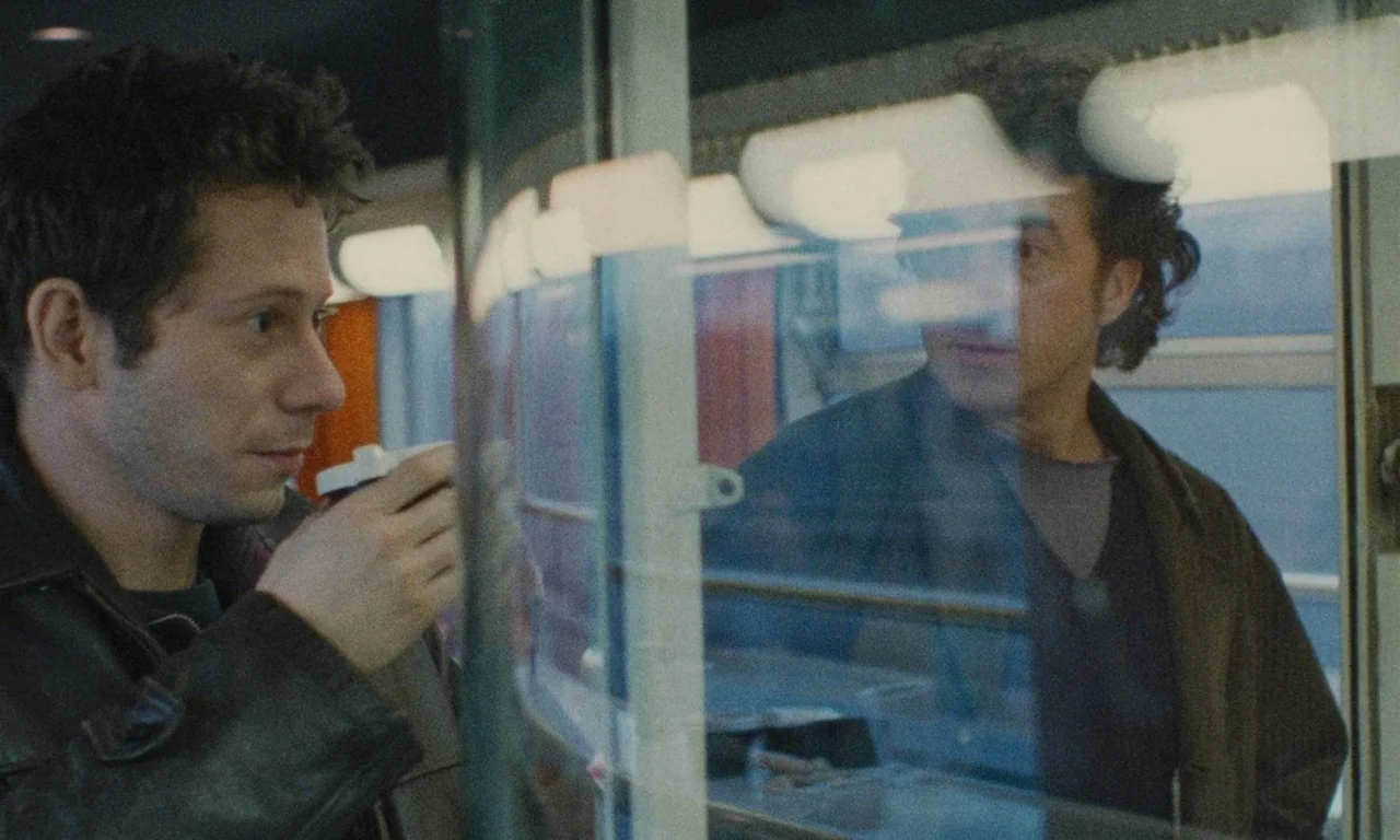 Late August, Early September (1998) movie still: medium — A modern train car interior with brushed metal walls and reflective glass partitions. A…