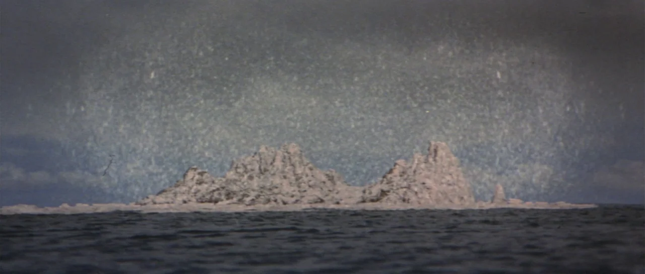 Son of Godzilla (1967) movie still: establishing — A craggy, snow-covered ice formation floats in dark, rippled ocean waters. Dense, gloomy clouds…