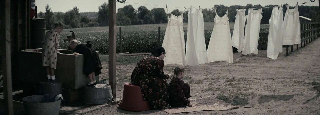 Women Talking (2022) movie still: wide — A rural farmyard features a row of white linens drying on a wire line. Several people in conservative…