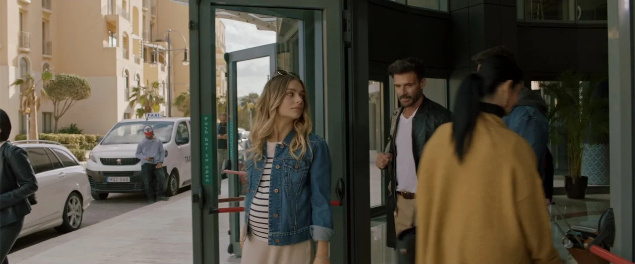 Hounds of War (2024) movie still: medium — A glass doorway connects a bright street to a modern building lobby interior. A pregnant woman in a denim…