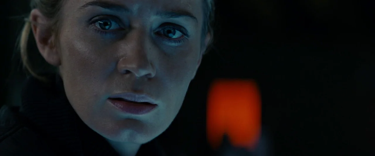 Edge of Tomorrow (2014) movie still: closeup — A woman's face is illuminated in a very dark room with black surroundings. A small, out-of-focus,…