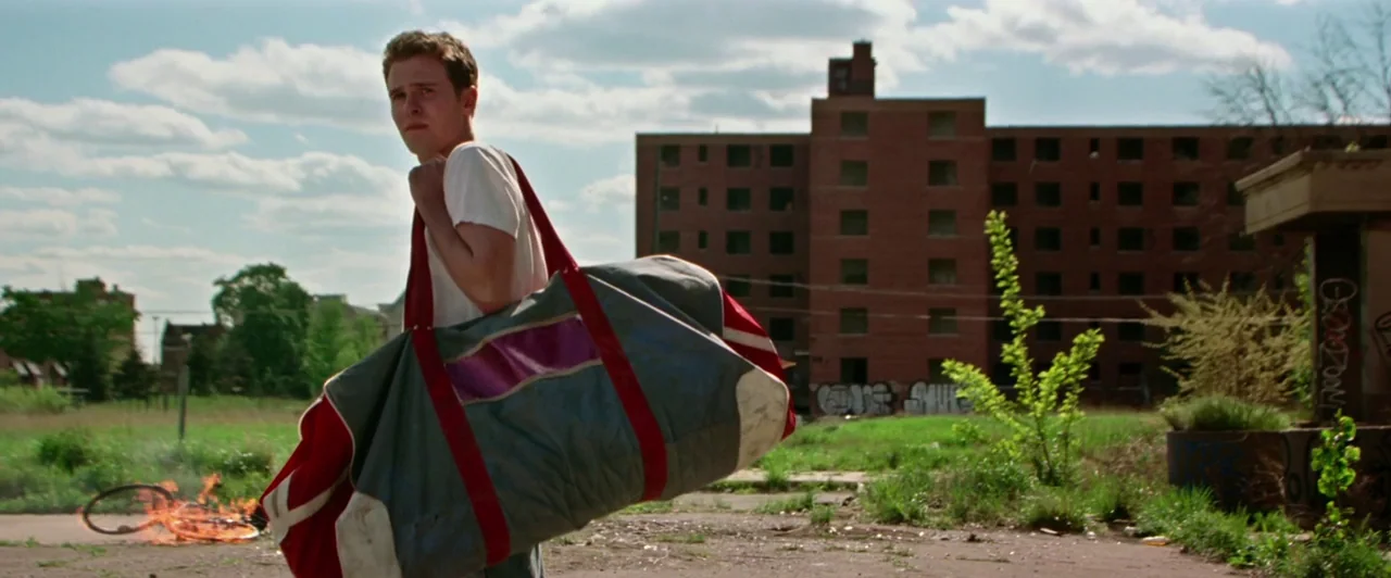 Lost River (2014) movie still: fullbody — A man walks past a burning bicycle in a blighted urban lot. A multi-story brick building stands in the…