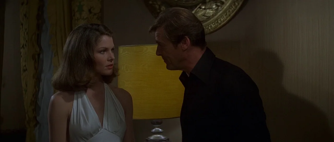 Moonraker (1979) movie still: medium — A dimly lit room features a glowing yellow table lamp and gold wall decor. A man in a dark shirt faces a…