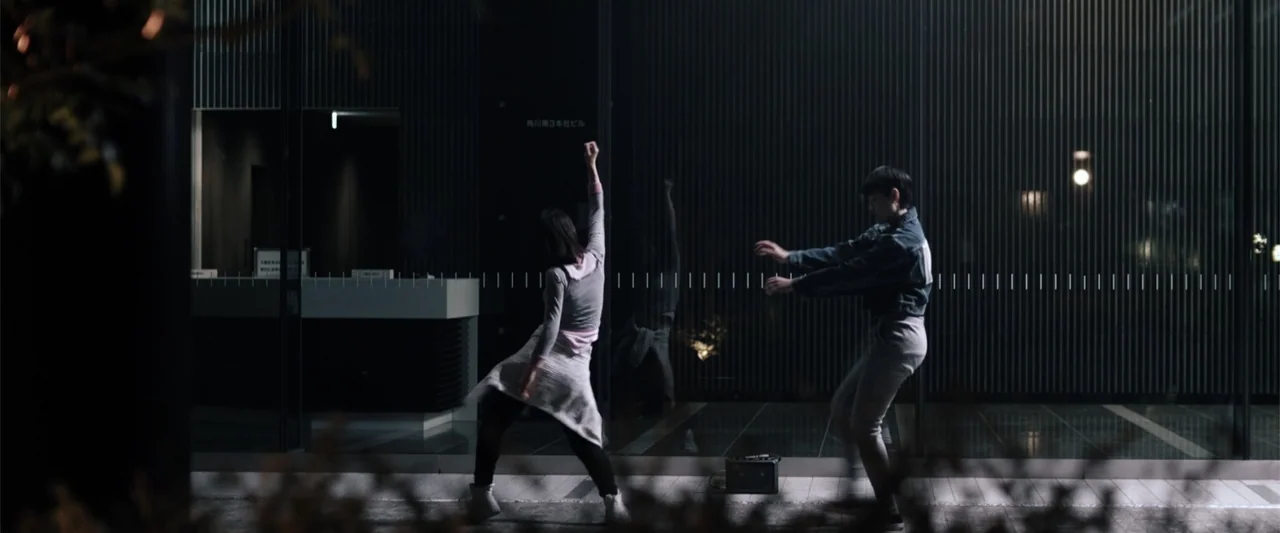To Hear the Birds Singing (1995) movie still: fullbody — A modern building with a dark glass wall stands at night. Two young people dance on the…