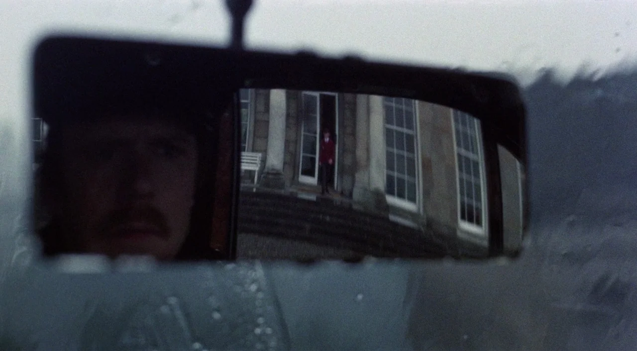 The Iguana with the Tongue of Fire (1971) movie still: closeup — A close view through a rearview mirror shows a man in a vehicle. A distant figure…