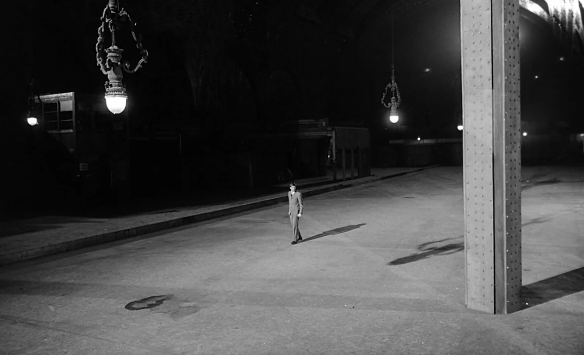 The Trial (1962) movie still: wide — A dark, expansive train station platform features large metal support columns and vaulted ceilings. A lone man…