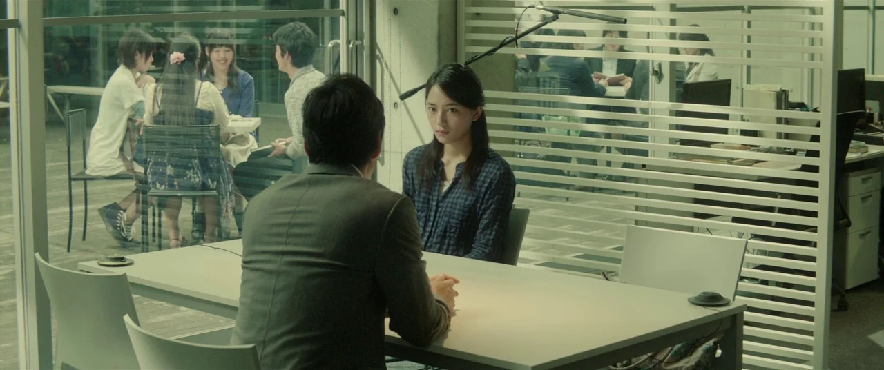 Creepy (2016) movie still: medium — A modern, sterile office meeting room with concrete walls and horizontal window blinds. A man in a suit faces a…