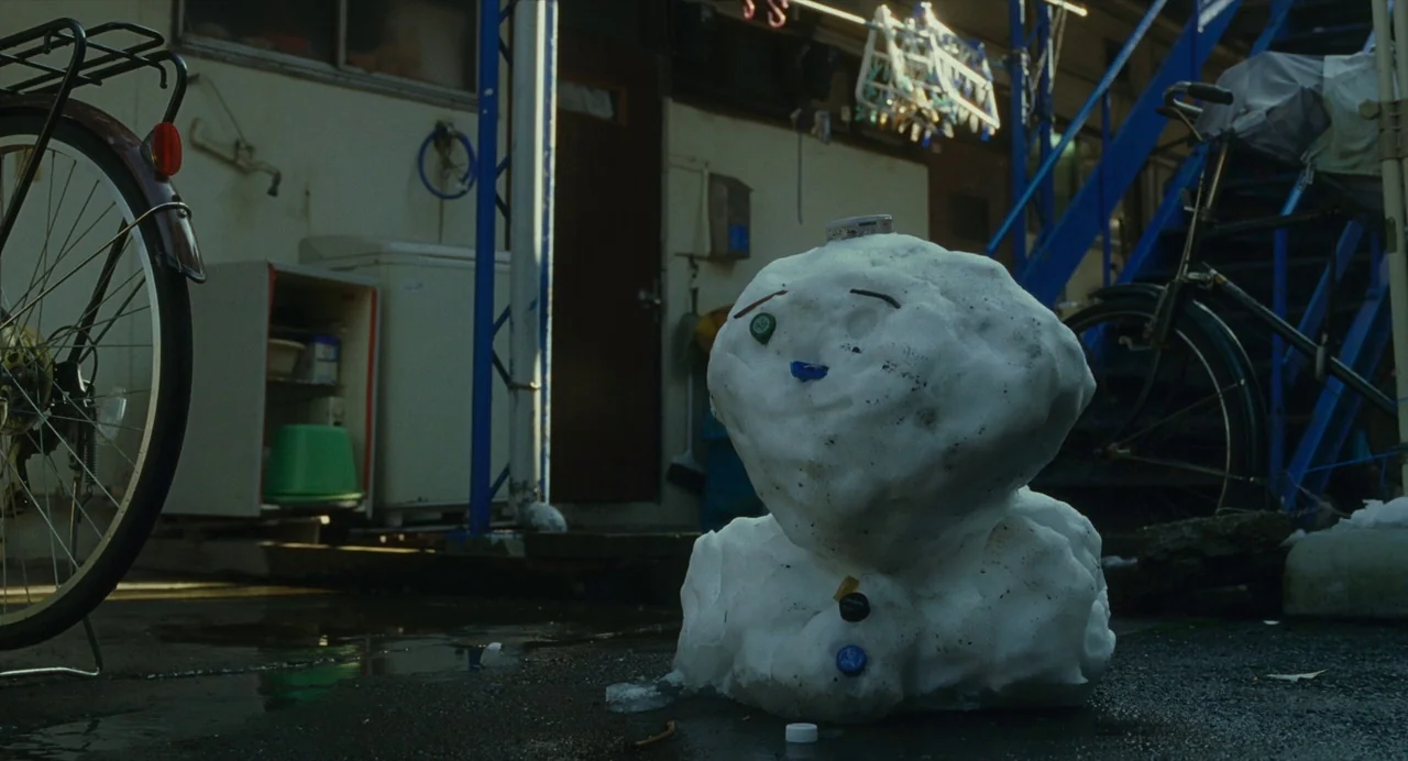 Shoplifters (2018) movie still: medium — A snow sculpture sits on wet asphalt in an apartment courtyard during winter. Bicycles and household items…