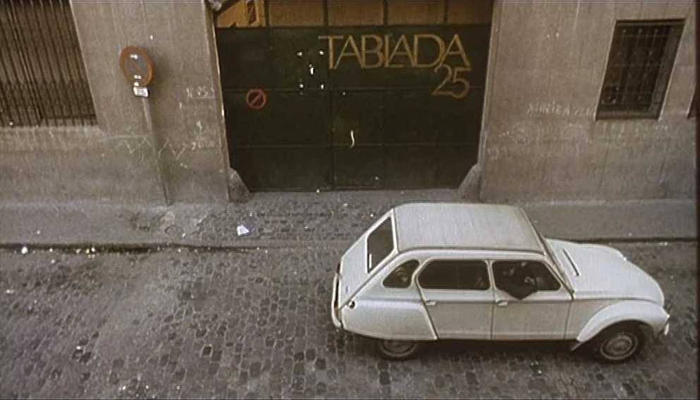 Labyrinth of Passion (1982) movie still: wide — A quiet cobblestone street features a large metal gate with gold lettering. A white vintage…