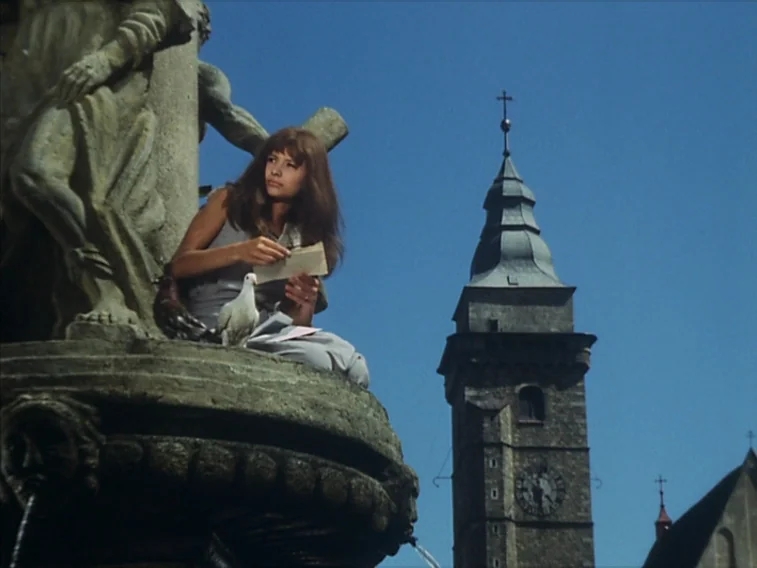 Valerie & Her Week of Wonders (1970) movie still: medium — A woman sits atop a large, ornate stone fountain in a sunny town square. A tall stone…