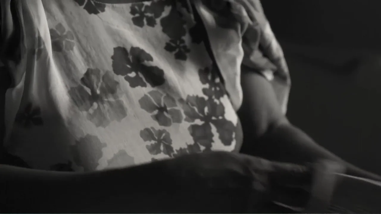 Faya Dayi (2021) movie still: medium — An interior space displays a person wearing a floral print top in dim light. The person sits while actively…