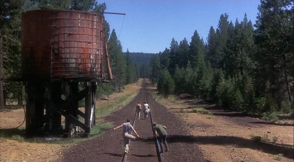Stand By Me (1986) movie still: wide — A remote set of railroad tracks runs through a dense forest with tall pines. Four teenage boys walk down the…