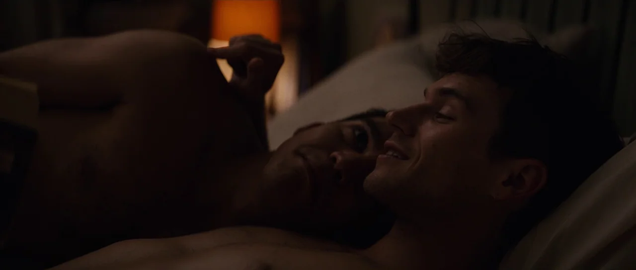 Hellraiser (2022) movie still: closeup — A dimly lit room with an out-of-focus orange lamp glow in the background. Two shirtless men lie in bed,…