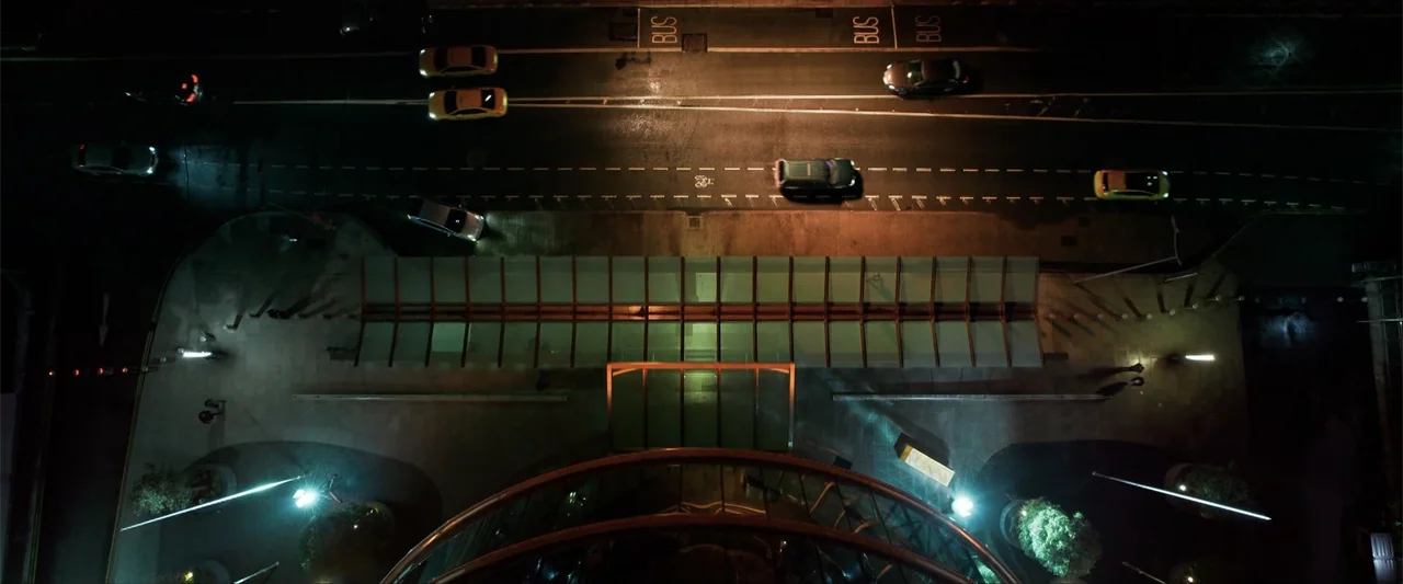 Drop (2025) movie still: establishing — A top-down view shows a glass-roofed bus stop on a dark street at night. Multiple cars travel along the…