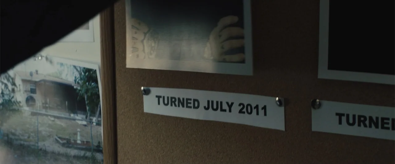 Skin (2009) movie still: closeup — A dark cork board displays various printed photographs and white paper labels with text. The labels indicate…
