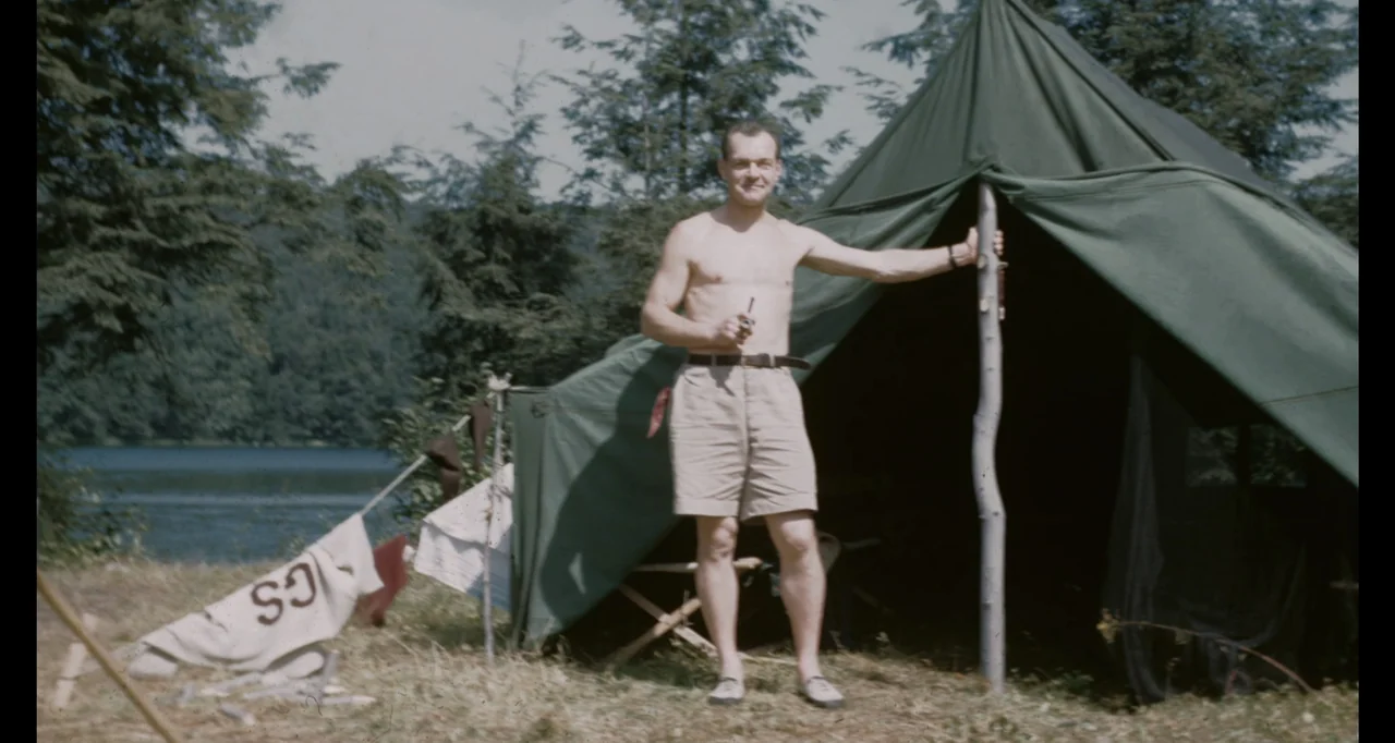 Living With Lincoln (2015) movie still: fullbody — A man stands before a green canvas tent located near a lakeside clearing. The subject wears khaki…