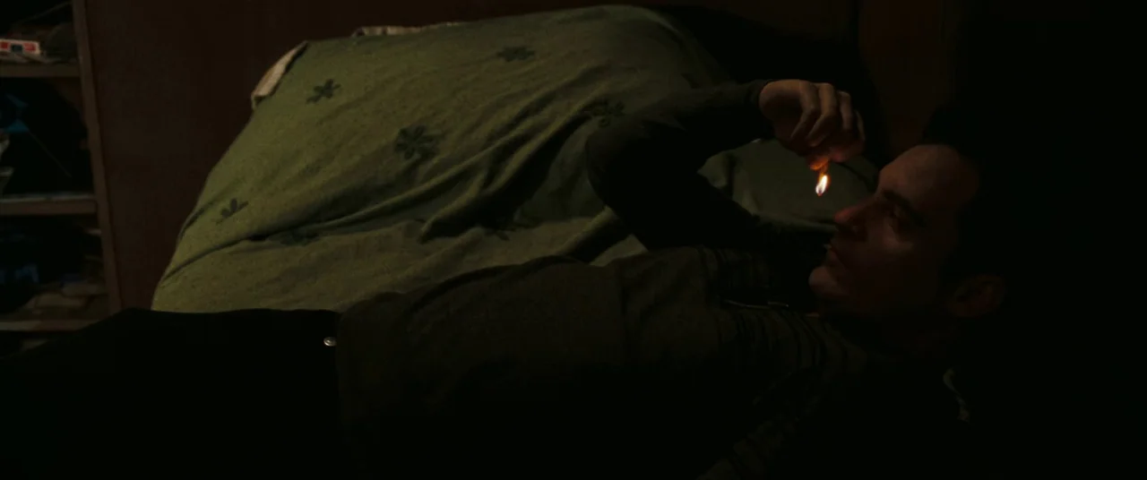 Two Lovers (2008) movie still: medium — A dark, dim room with a green bedspread featuring a small flower pattern. A man lies on his back on the bed…