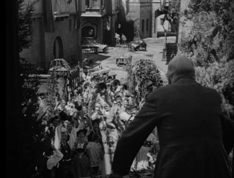 Frankenstein movie still: wide — A crowded European-style town square with people gathered to celebrate in the sunshine. An older man in a suit…