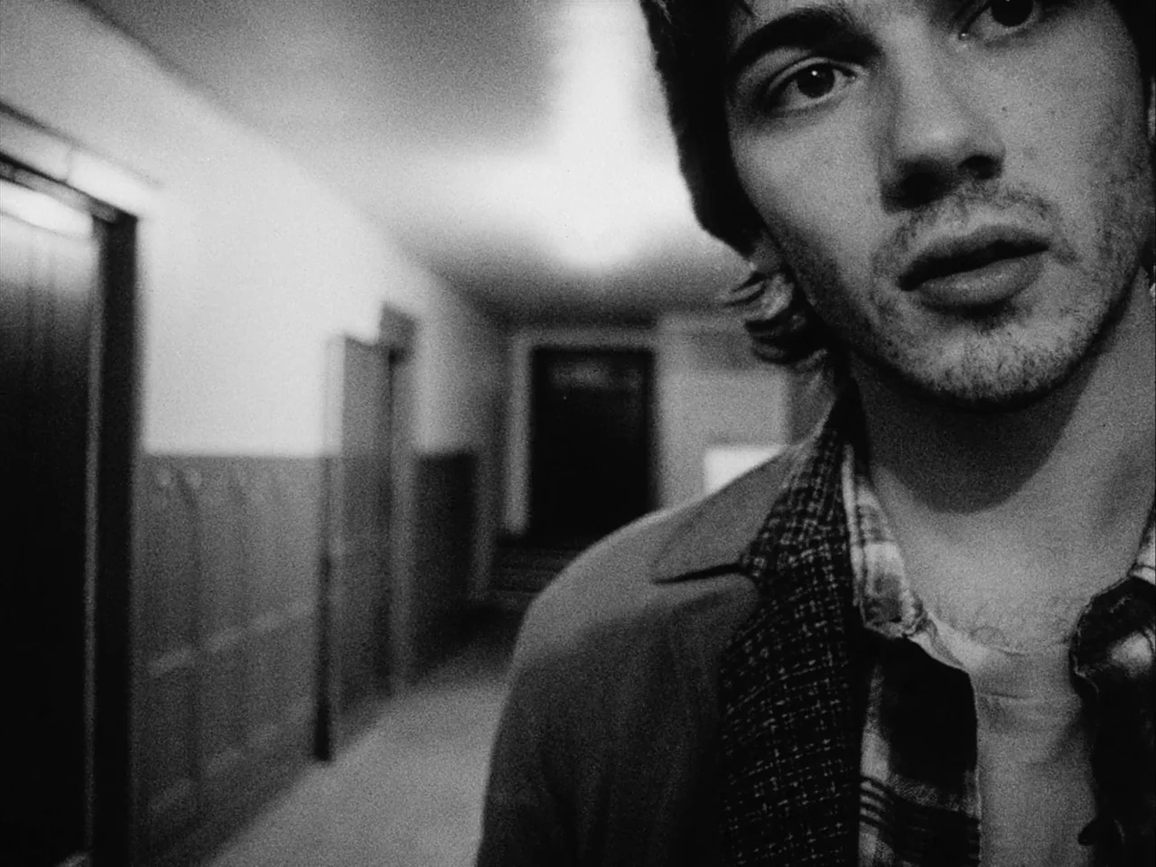 Mala Noche (1986) movie still: closeup — A dimly lit indoor corridor with wooden wainscoting and doors along the wall. A young man stands in the…