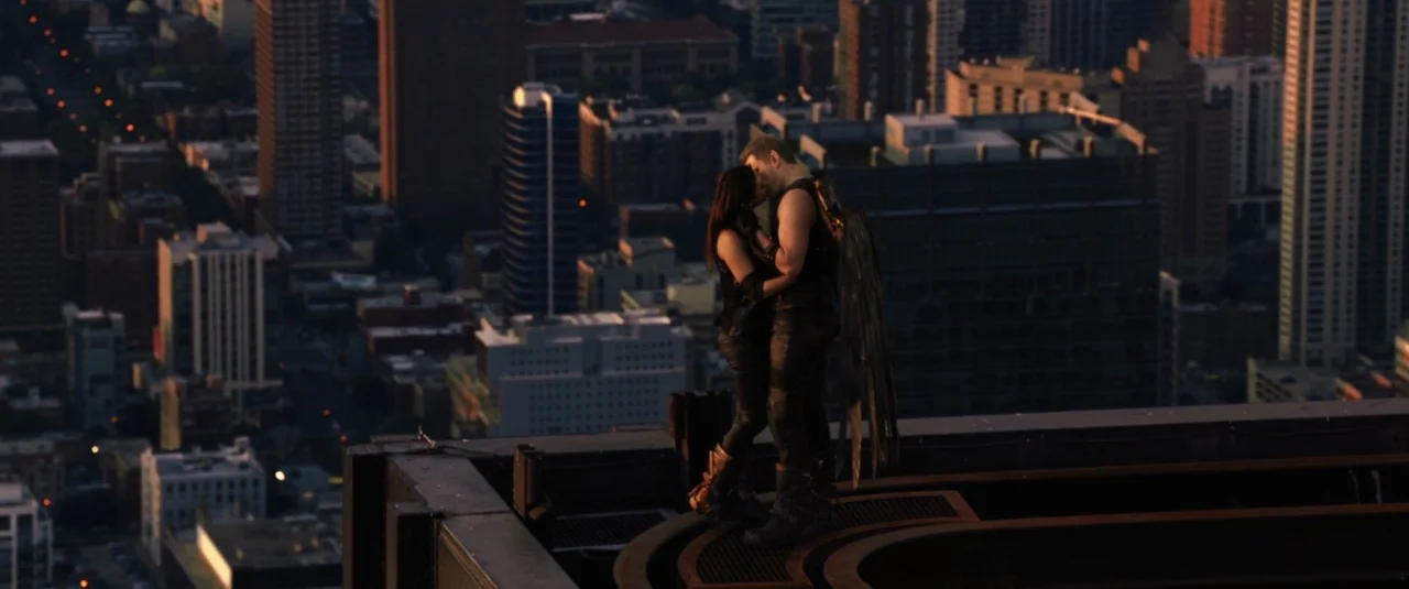 Jupiter Ascending (2015) movie still: wide — A man and a woman stand atop a high building overlooking a city at sunset. The man has large wings…