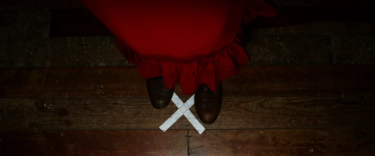 Pearl (2022) movie still: closeup — A dark wooden floor features a white tape mark in the shape of an X. A person wearing a long red skirt stands…