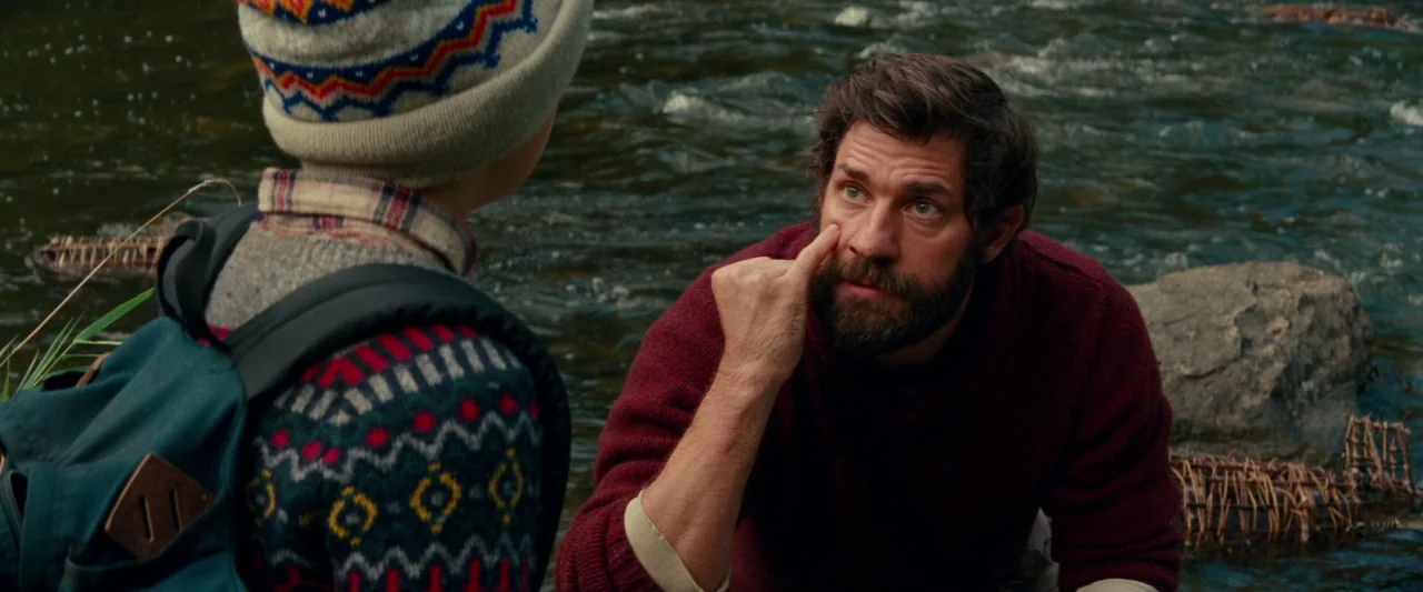 A Quiet Place (2018) movie still: medium — A man and a child crouch beside a flowing river with small wicker traps. The man touches his face while…