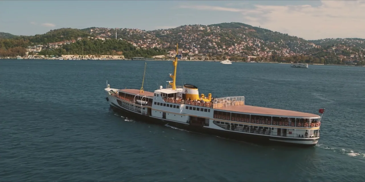 Kill Me If You Dare (2019) movie still: wide — A passenger ferry cruises across a large body of water near a coastal city. The ship is white and…