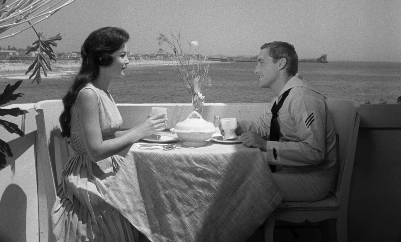 Night Tide (1961) movie still: medium — A sunny balcony overlooking the ocean with a small table for two guests. A young woman and a sailor in…