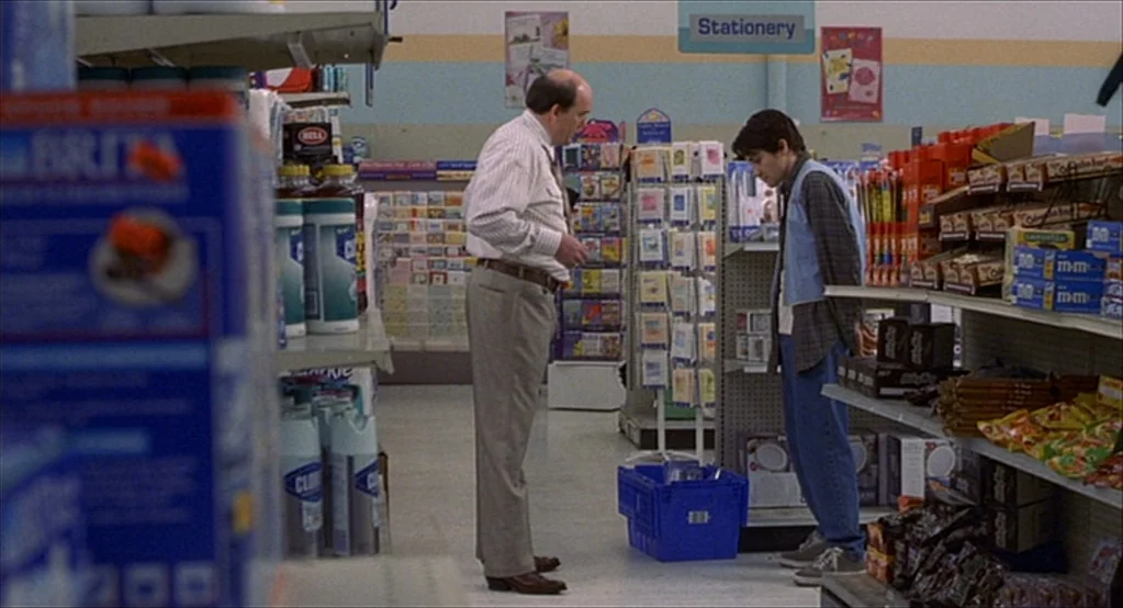 The Good Girl (2002) movie still: fullbody — An aisle in a brightly lit retail store features rows of products and greeting cards. An older man…