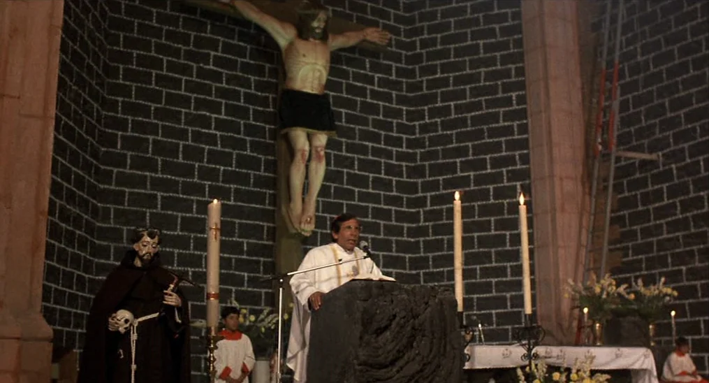 Salvador (1986) movie still: wide — A dark stone church interior features a large wooden crucifix mounted on a wall. A priest speaks from a dark…