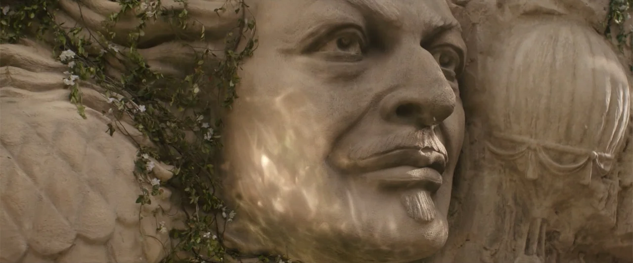 Wicked (1998) movie still: closeup — A stone relief sculpture is integrated into a wall surrounded by small green vines. Sunlight reflecting from…