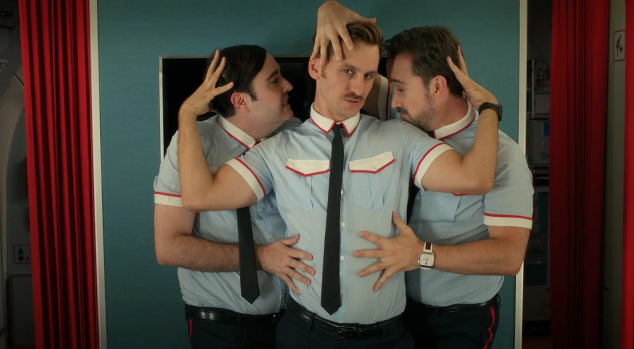 I’m So Excited! (2013) movie still: medium — Three men in matching airline uniforms stand together in an airplane galley. They are positioned…