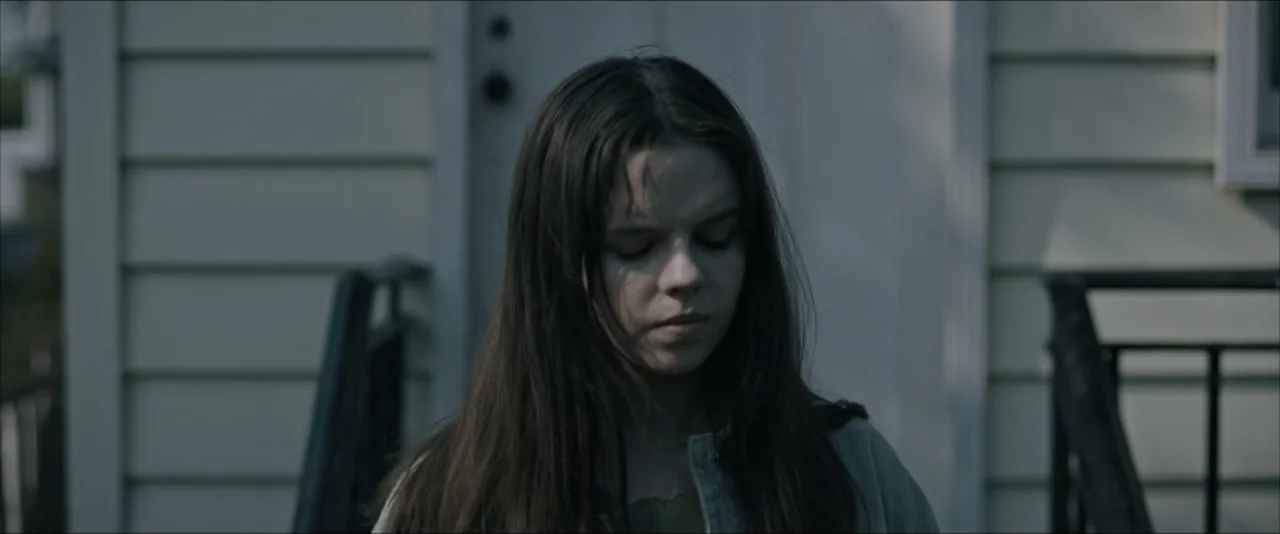 It Feeds (2025) movie still: medium — A young person stands on the porch of a residential house with light siding. The subject is looking downward…