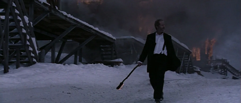 The Claim (2000) movie still: fullbody — A snowy, desolate encampment features wooden structures partially obscured by dark, billowing smoke. A man…