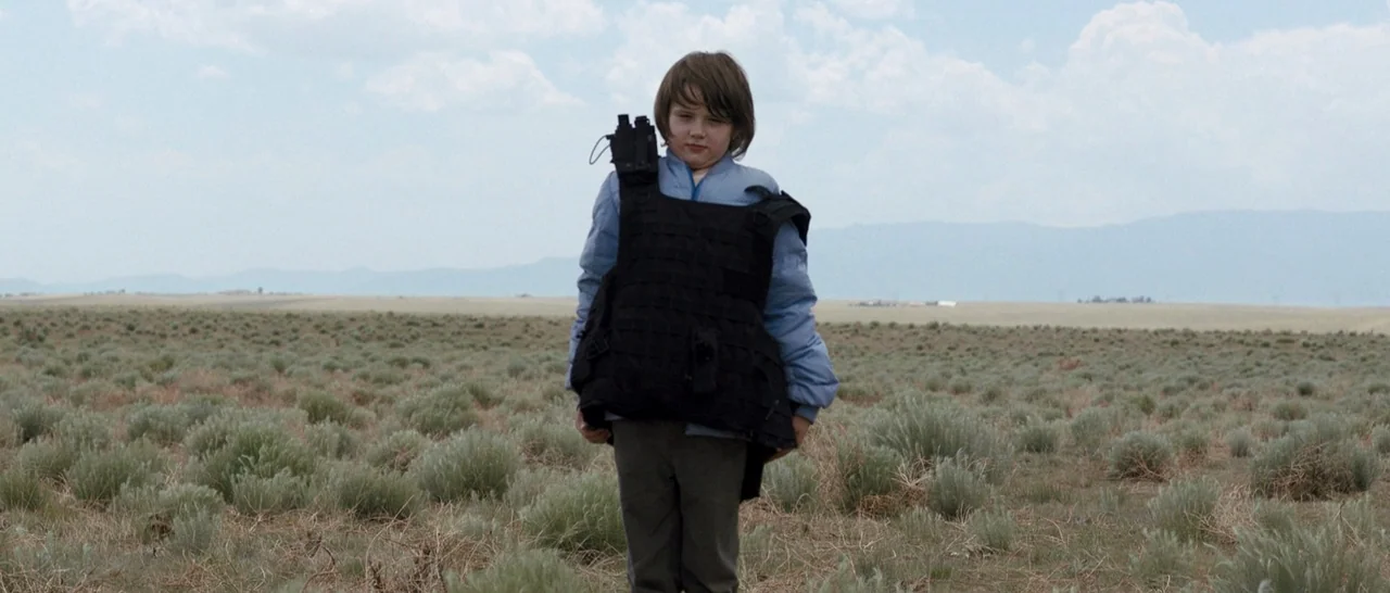 Cop Car (2015) movie still: fullbody — A vast, dry desert landscape with low-lying brush under a hazy, overcast sky. A young child wearing a…