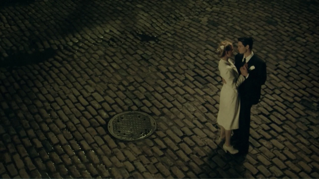 The Words (2012) movie still: wide — A nighttime cobblestone street features a manhole cover nearby. A couple in formal wear dances together in the…