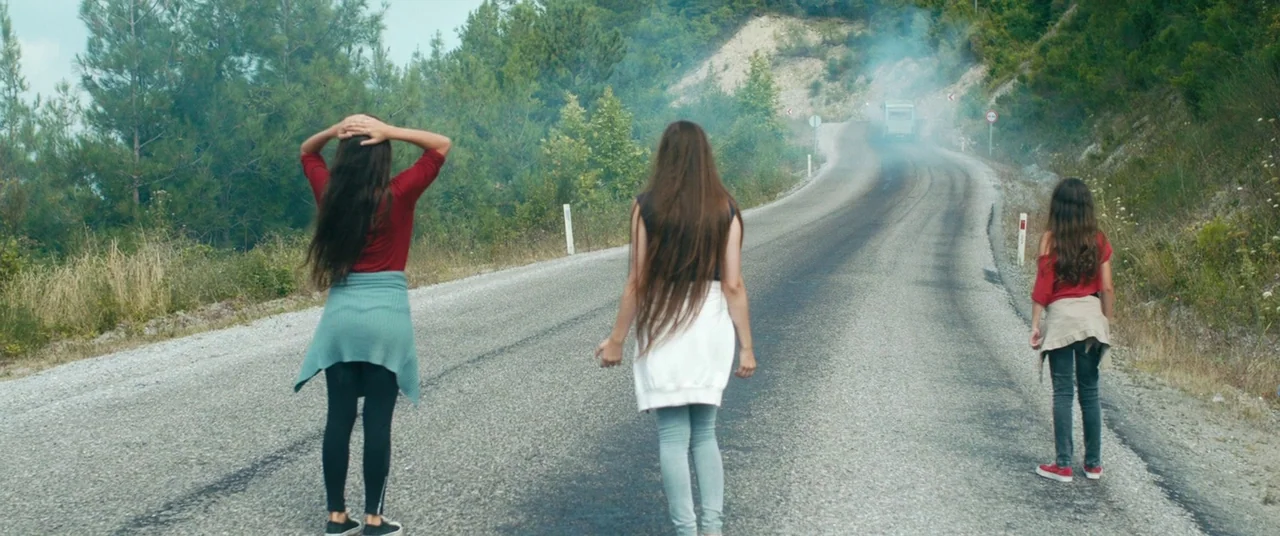 Mustang (2015) movie still: fullbody — Three young girls walk away from the camera on a paved mountain road. A plume of white smoke rises from a…